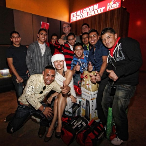 A group of people, including a woman in a Santa hat, posing with gifts under a "Jenni's Noodle House" sign.