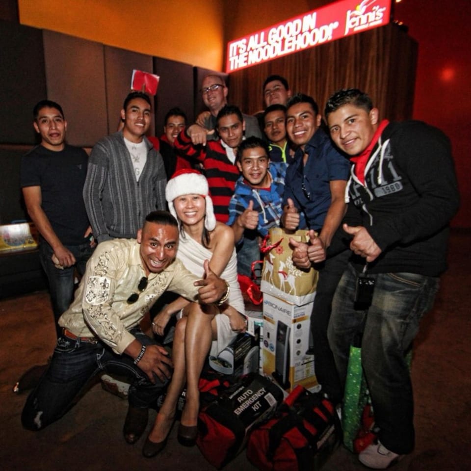 A group of people, including a woman in a Santa hat, posing with gifts under a "Jenni's Noodle House" sign.