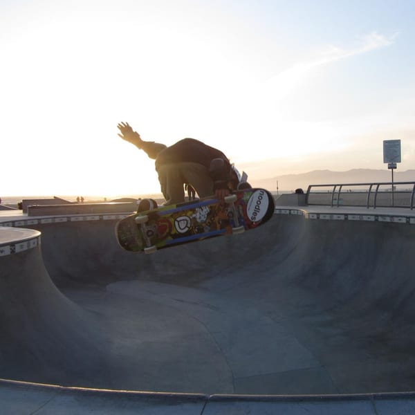 Skateboarder in mid-air trick over a concrete skate park bowl at sunset, with ocean and mountains in background.