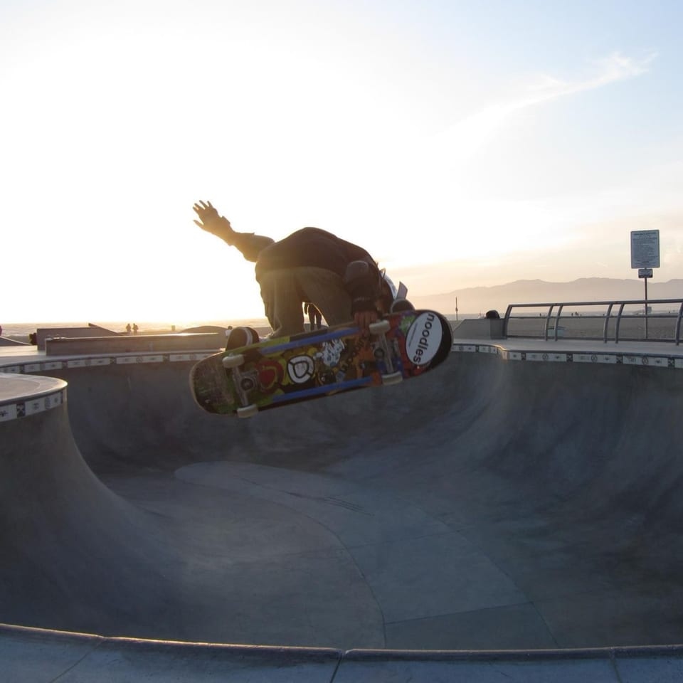 Skateboarder in mid-air trick over a concrete skate park bowl at sunset, with ocean and mountains in background.